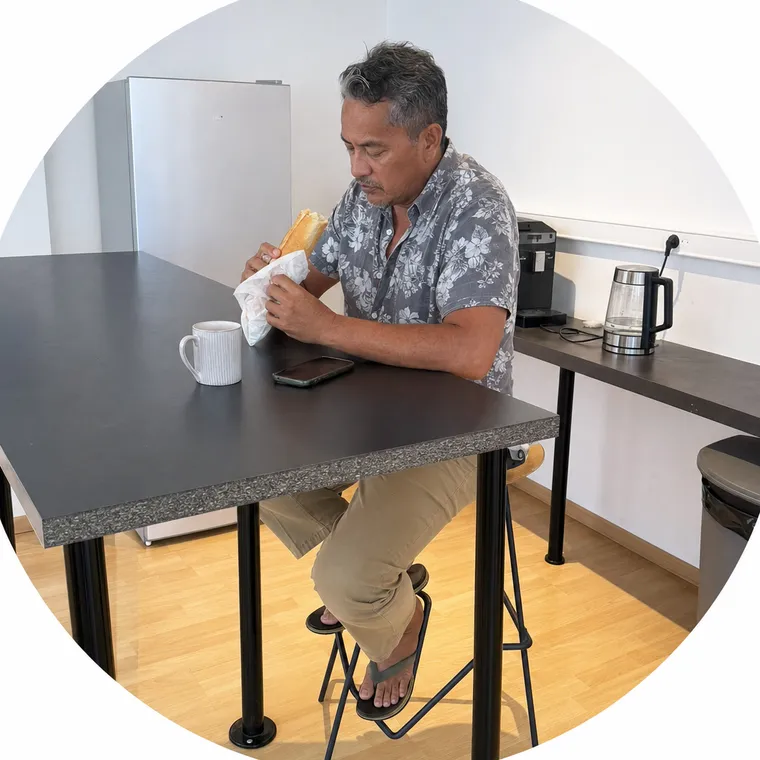 Un homme déjeune d'un sandwich, assis à la table haute de la cafétéria, un café posé devant lui