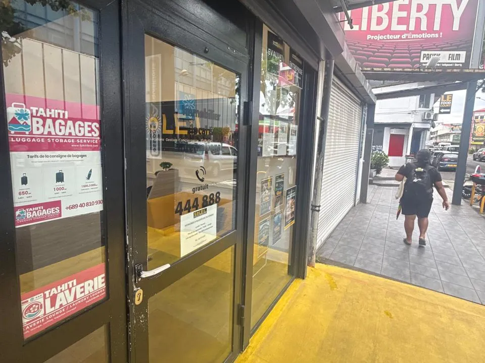 L'entrée sous la galerie au sol jaune : une porte vitrée noire, le logo Tahiti Laverie collé sur la vitrine, l'enseigne du cinéma Liberty juste à côté